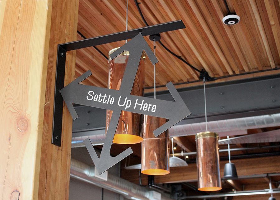 A crux-shaped sign above the counter reads, “settle up here”  the nail-laminated timber ceiling is clearly visible behind